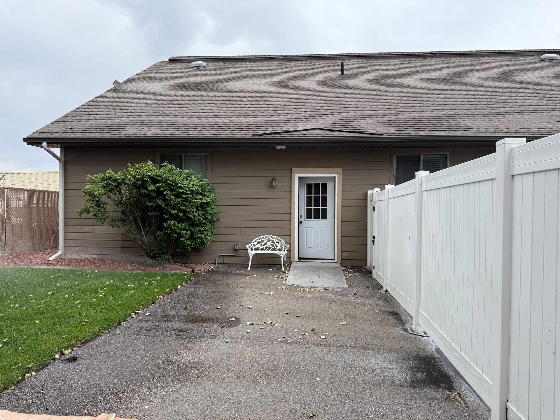 Back of a brown house with white door, a white bench, and a white fence; gravel path and green grass.