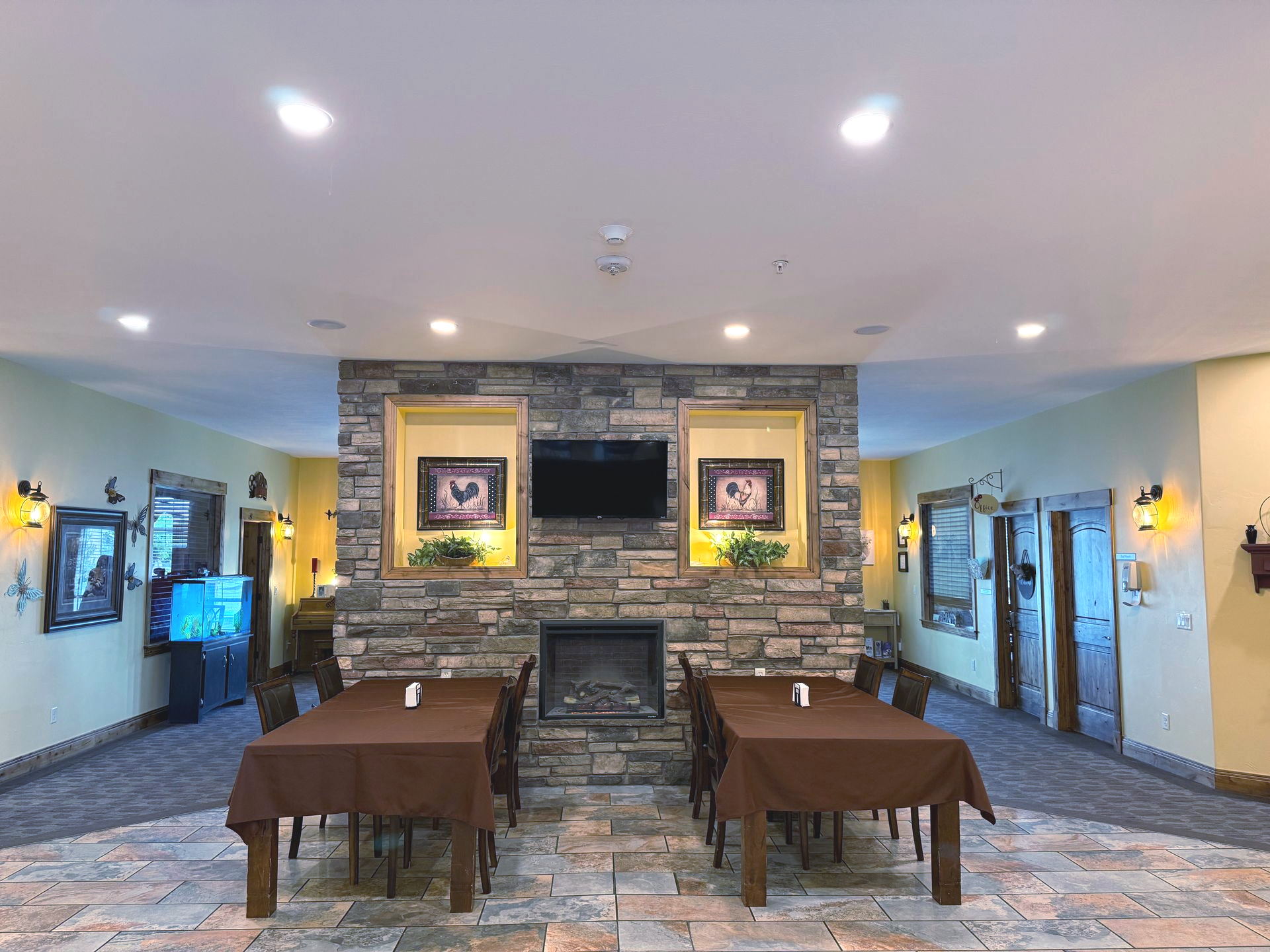 Dining area with stone fireplace, two tables with brown tablecloths, artwork, and recessed lighting.