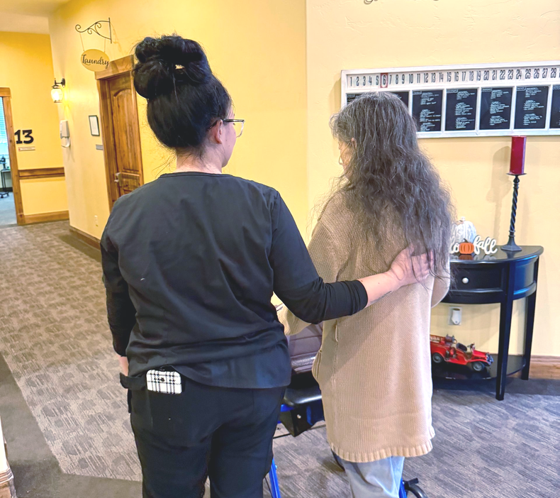 A caregiver in black scrubs assists an elderly woman with long gray hair, supporting her while she walks.