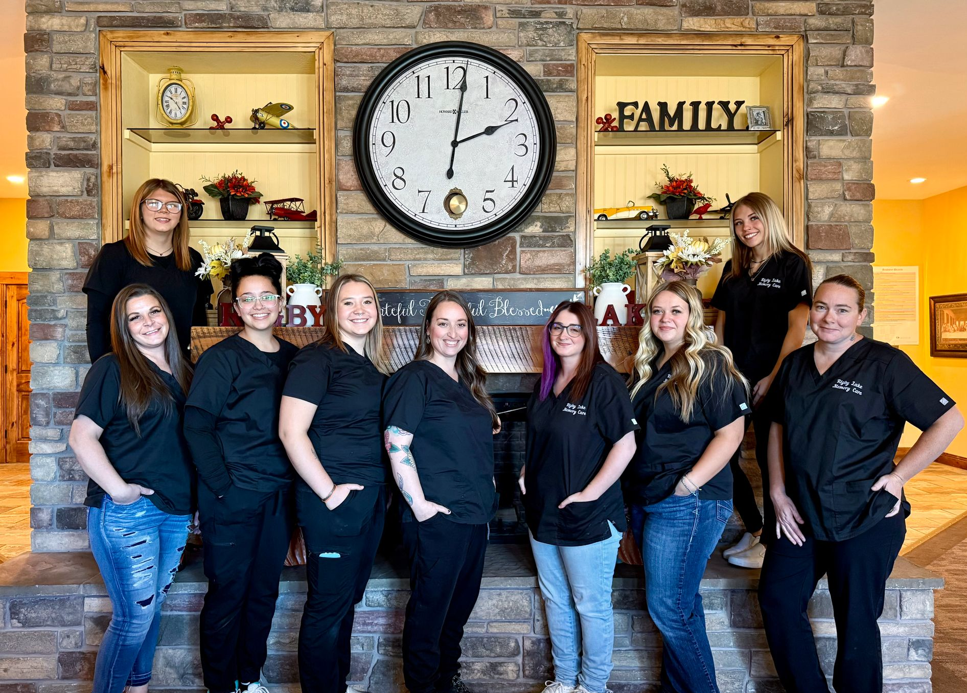 Group of dental staff in scrubs, smiling, standing in front of a brick fireplace with a large clock.
