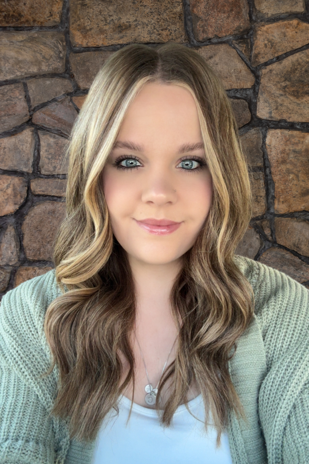 Woman with blonde highlights smiles, wears a green sweater and white top, against a stone wall.