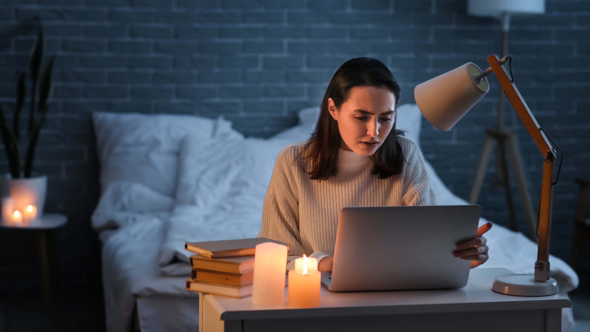 Woman working on a laptop at a desk in a dimly lit room with candles and a bed.
