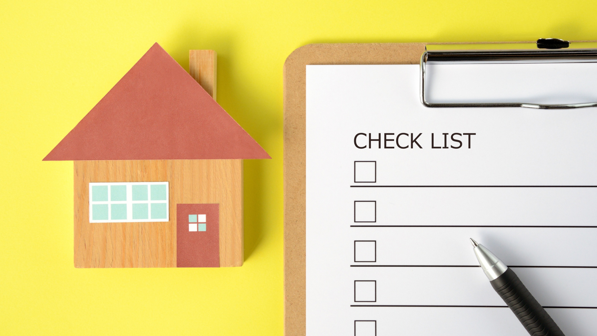 Wooden house model next to checklist on clipboard, pen, yellow background.