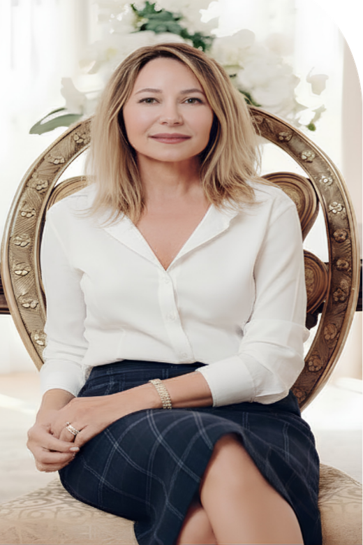 Woman in white outfit sitting in an ornate chair, smiling. Flowers and table in background.