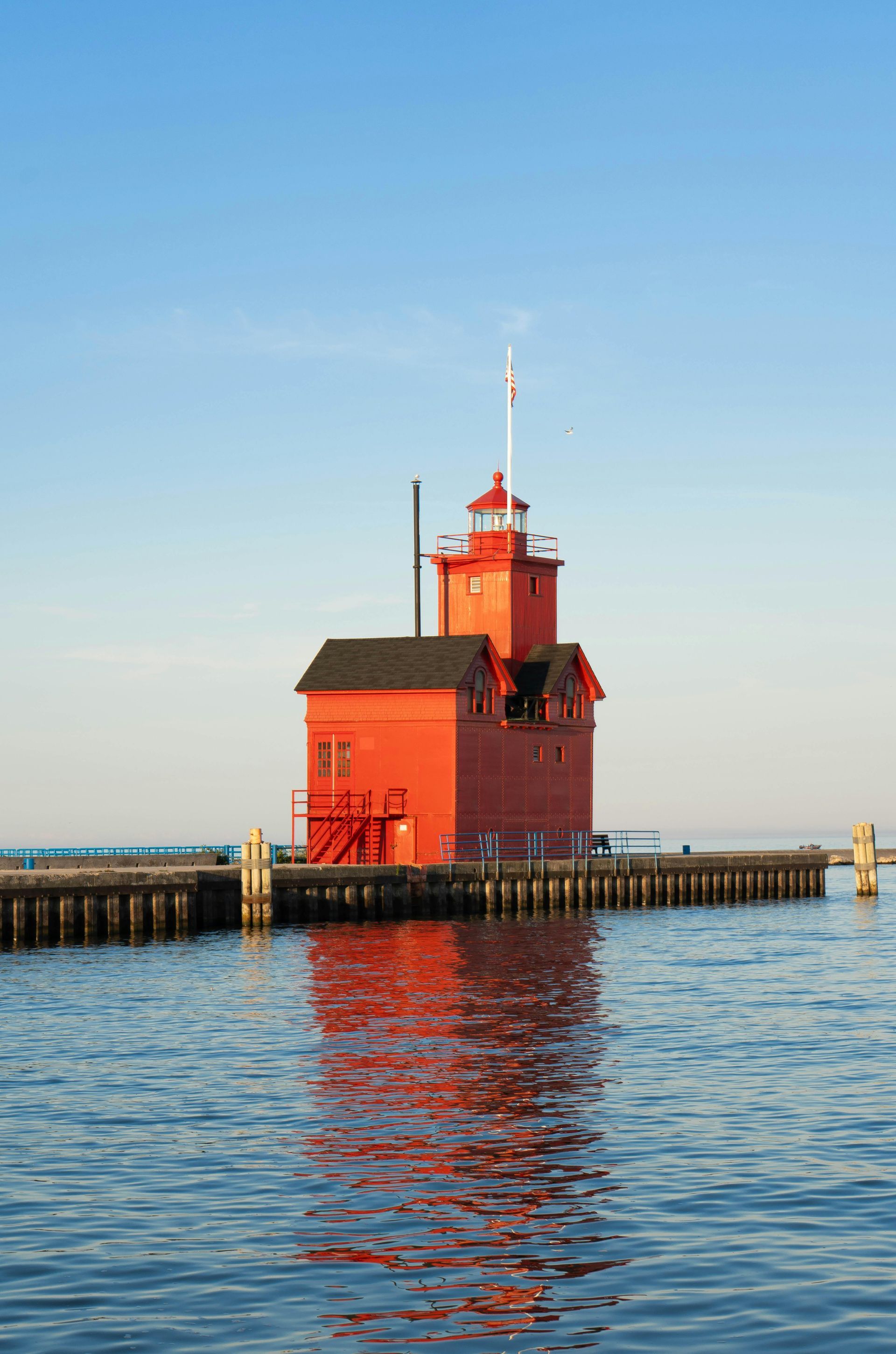Red lighthouse on pier, reflecting in calm water, blue sky.
