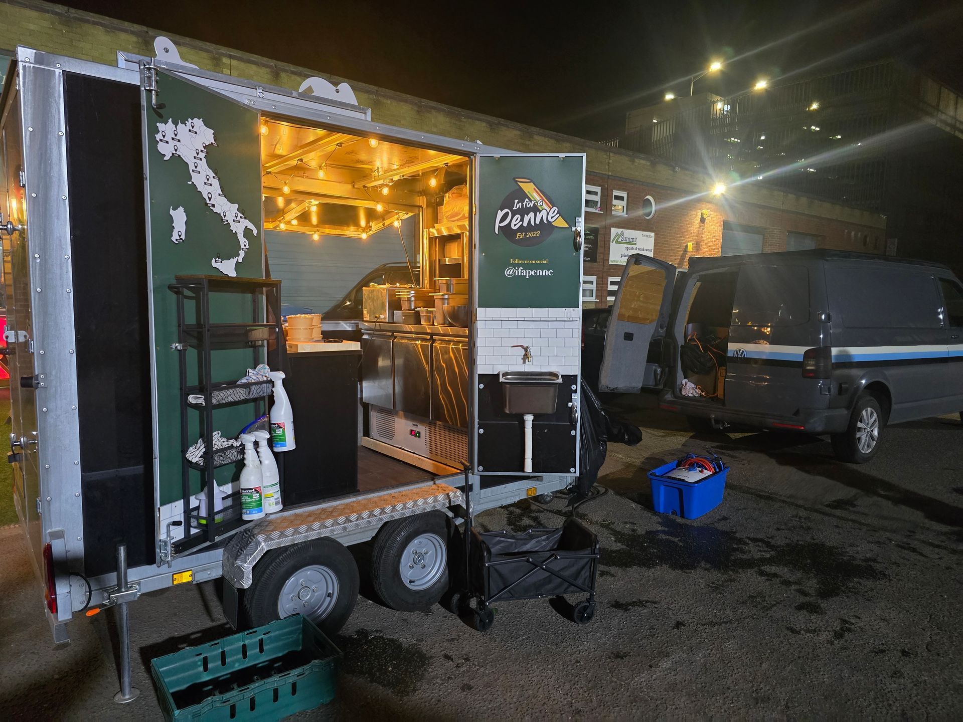 Lit food trailer at night beside a parked van, with cooking gear and equipment outside.