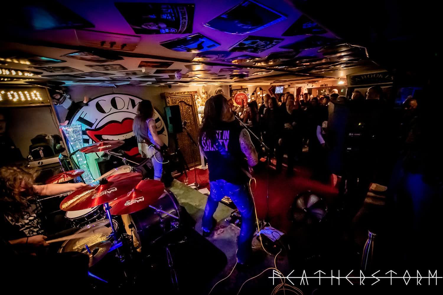 Live rock band performing in a dim club, with red drums, blue lighting, and a crowded stage.