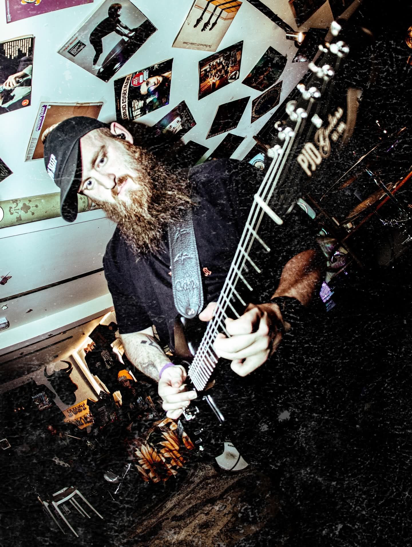 Long-haired guitarist performing indoors under posters, holding an electric guitar at a tilted angle.