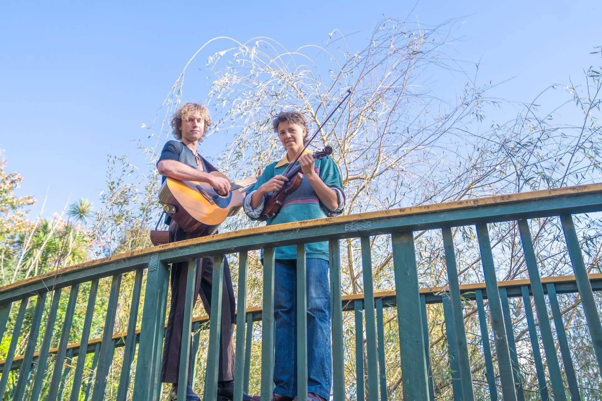 Two people stand behind a green railing, playing guitars outdoors under a blue sky.