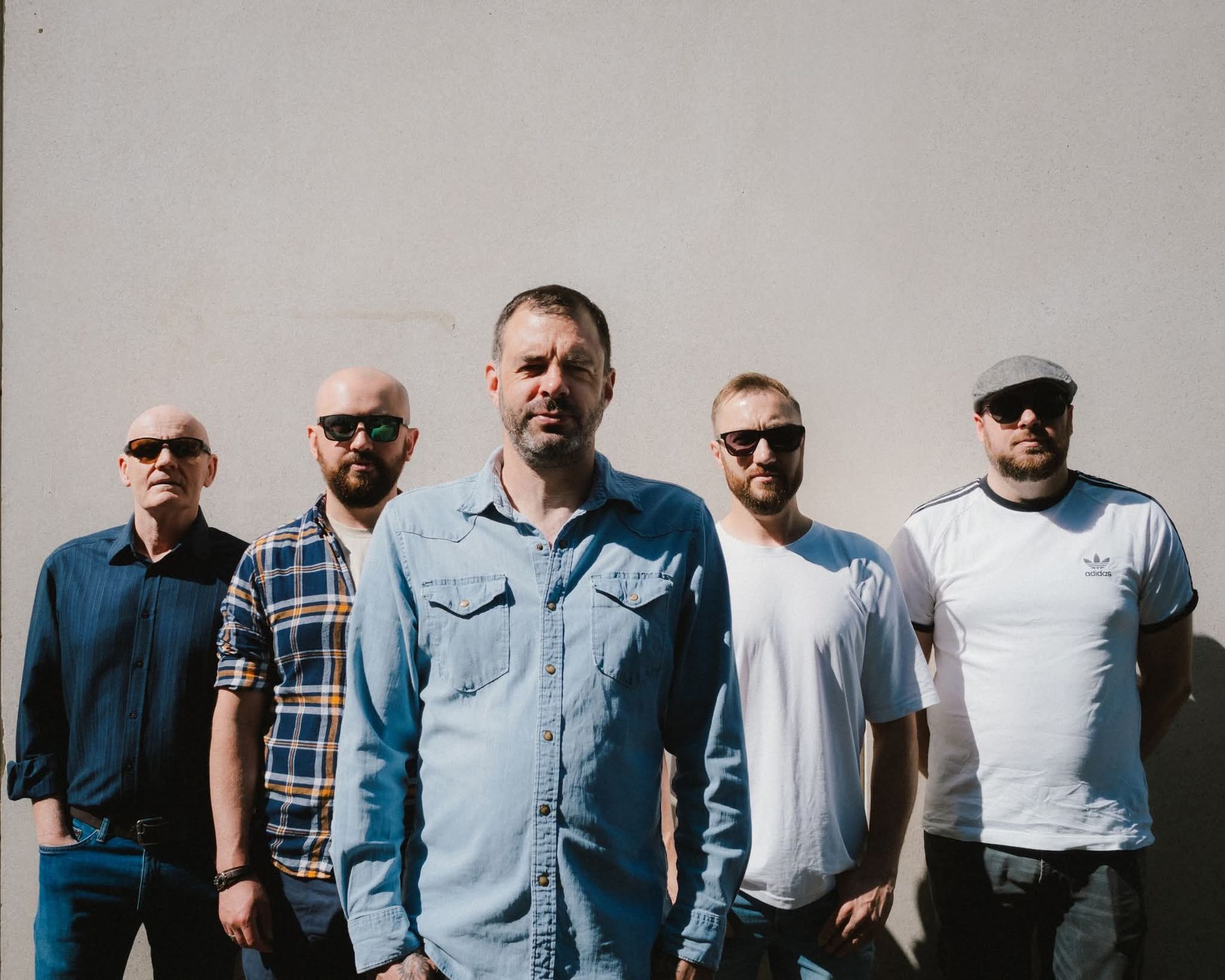 Five men standing outdoors against a blank wall, with two in sunglasses and one in a denim shirt in front