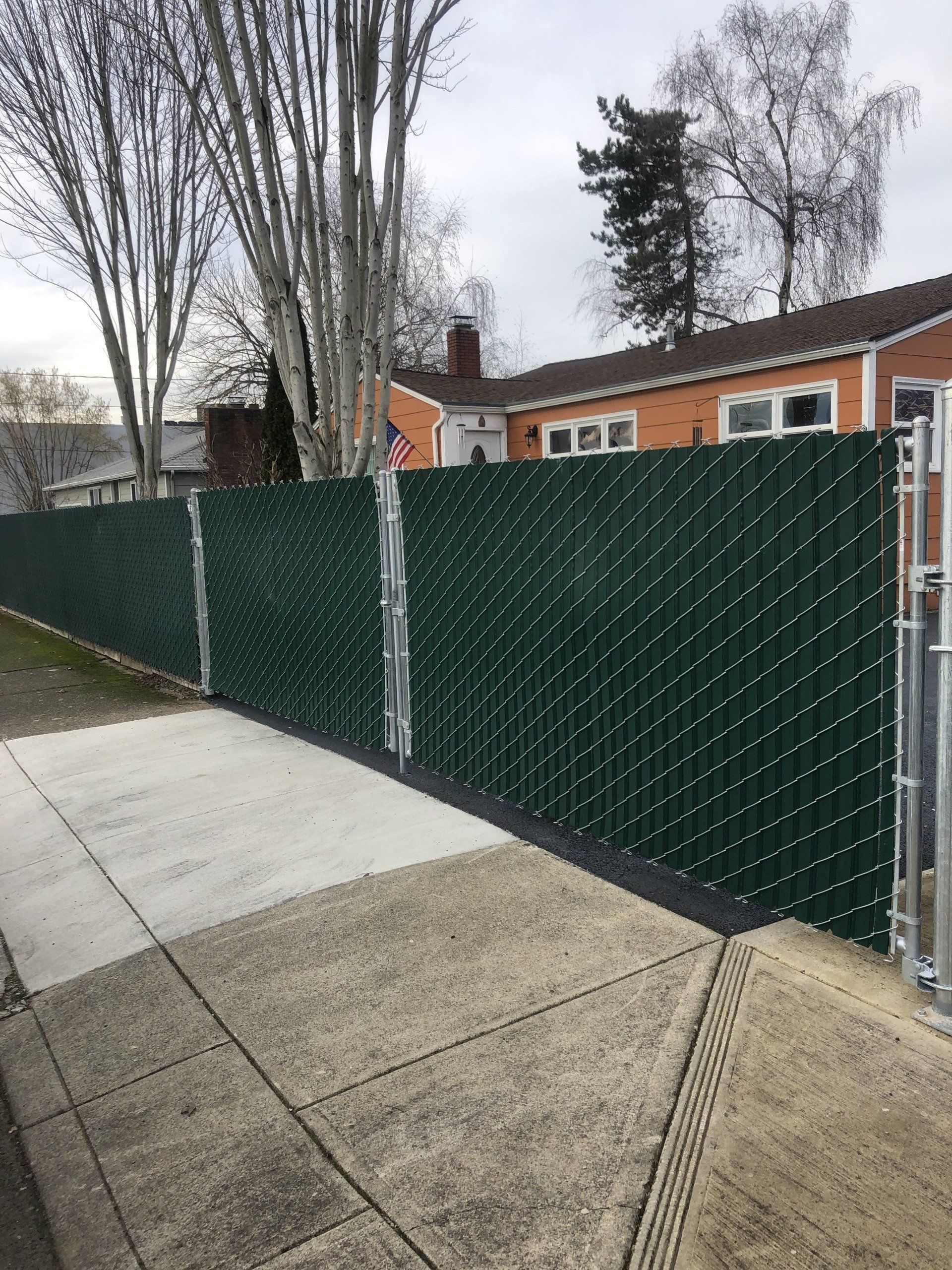 Green chain-link fence with privacy slats along a sidewalk. A house is in the background.