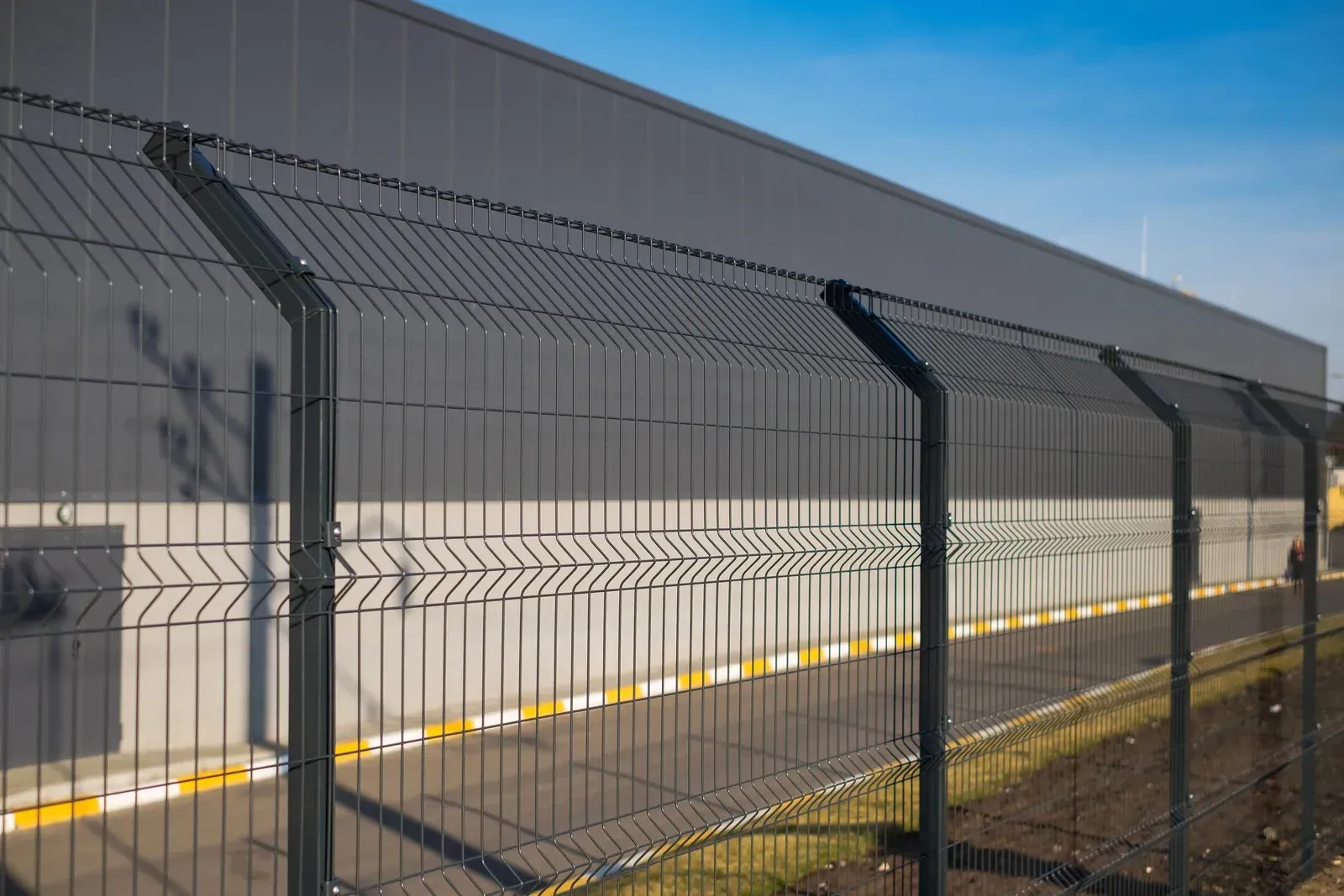 Dark gray wire fence in front of a large, gray building under a blue sky.