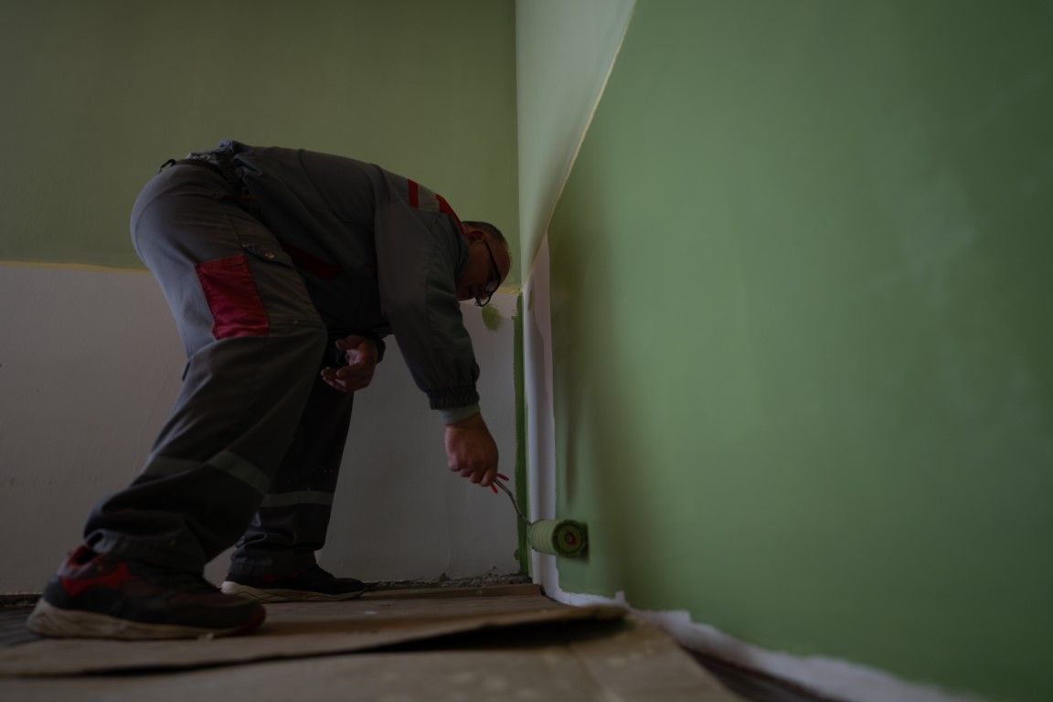 Person paints a green wall corner, holding a roller, in a room with white and green walls.