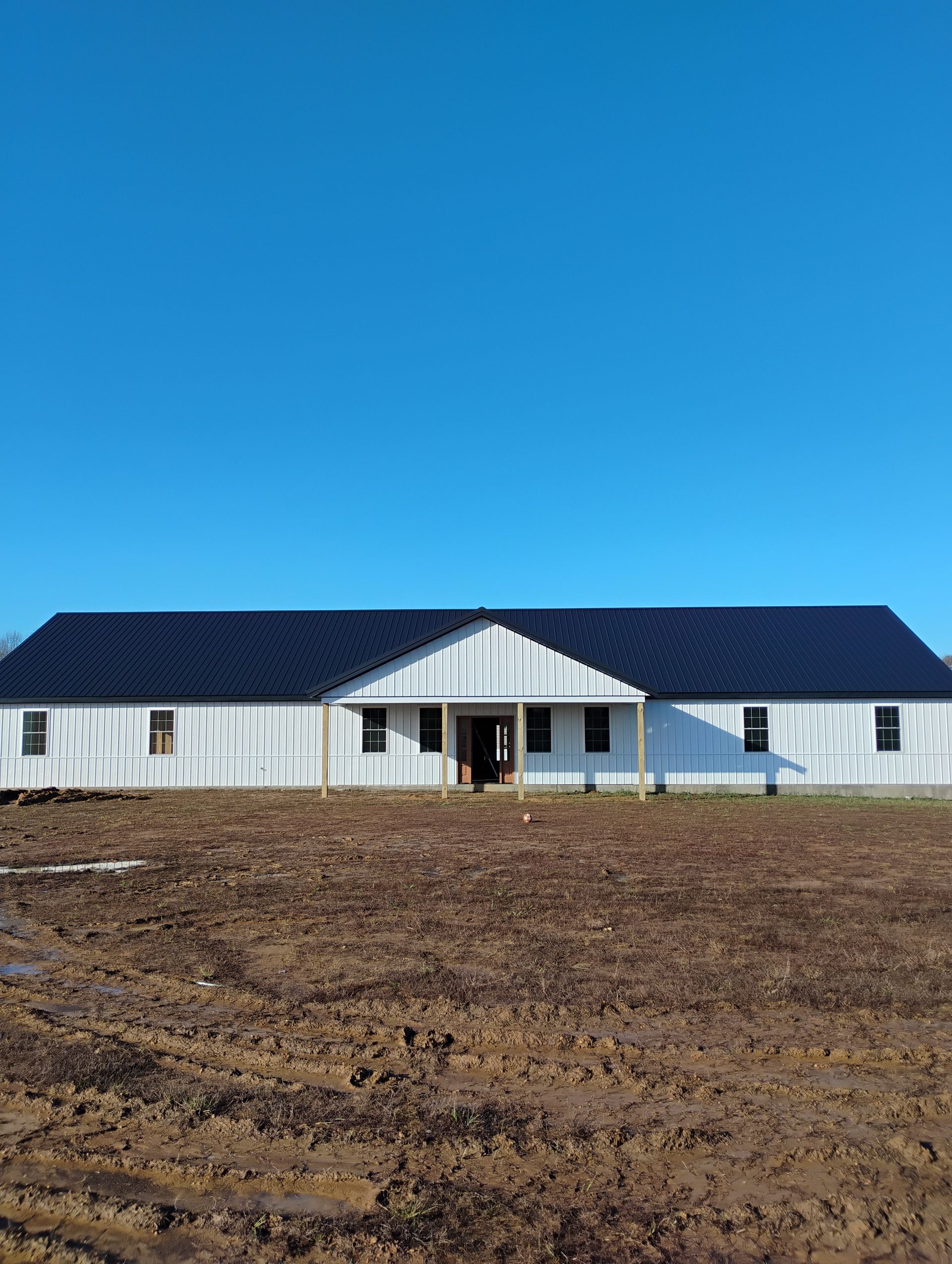 A large white house with a black roof is sitting in the middle of a dirt field.