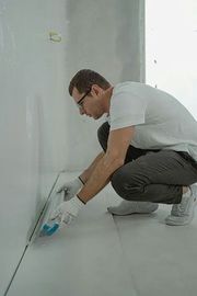 A person wearing white gloves and a white shirt crouches to smooth a white surface along a wall in an unfinished room.
