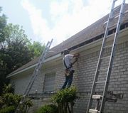 A construction worker stands on a ladder, installing or repairing the trim and fascia on the edge of a brick house roof.
