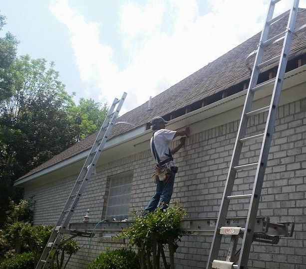 A construction worker stands on a ladder, installing or repairing the trim and fascia on the edge of a brick house roof.