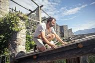 A man kneeling on a rustic wooden roof structure, placing boards in a partially constructed stone ruin.