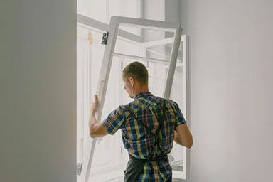 A person wearing a plaid shirt works to install or remove a white-framed window in a bright room.