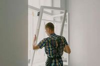 A person wearing a plaid shirt works to install or remove a white-framed window in a bright room.