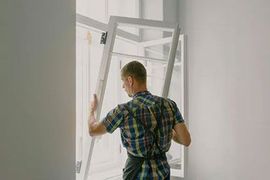A person in a plaid shirt installs a white window frame in a room with white walls.
