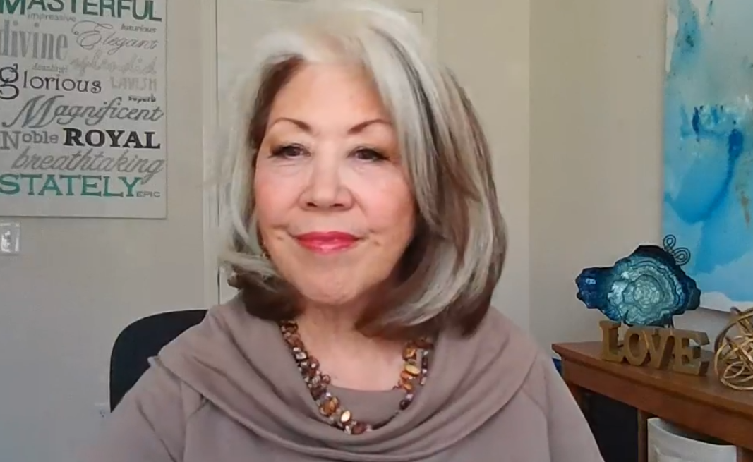 Woman with silver hair and mauve top, smiling indoors beside blue decor and framed wall art.