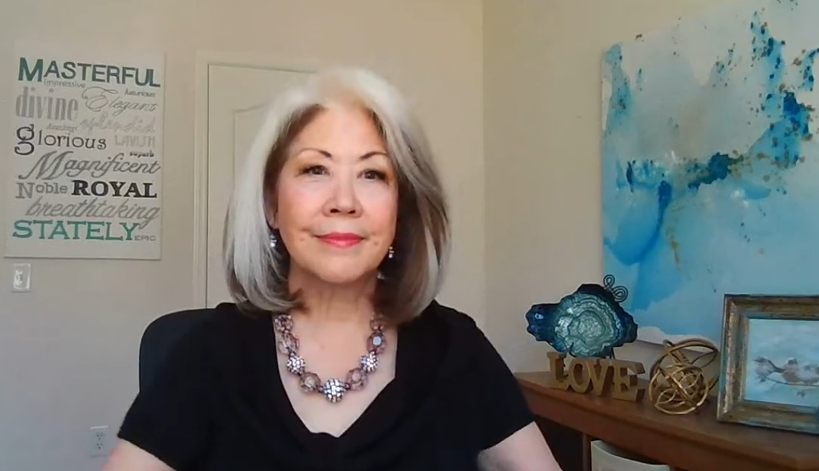 Woman seated indoors beside blue abstract art and decorative wall decor, wearing a black top and statement necklace.