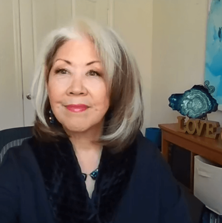Woman with silver hair and navy top, smiling indoors beside a shelf with a blue decorative plate and “LOVE” sign