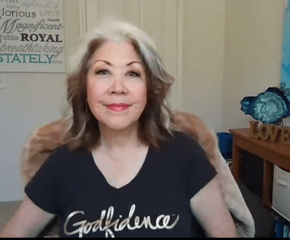 Woman seated indoors, smiling, wearing a black “Confidence” T-shirt, with a framed sign and decor behind her
