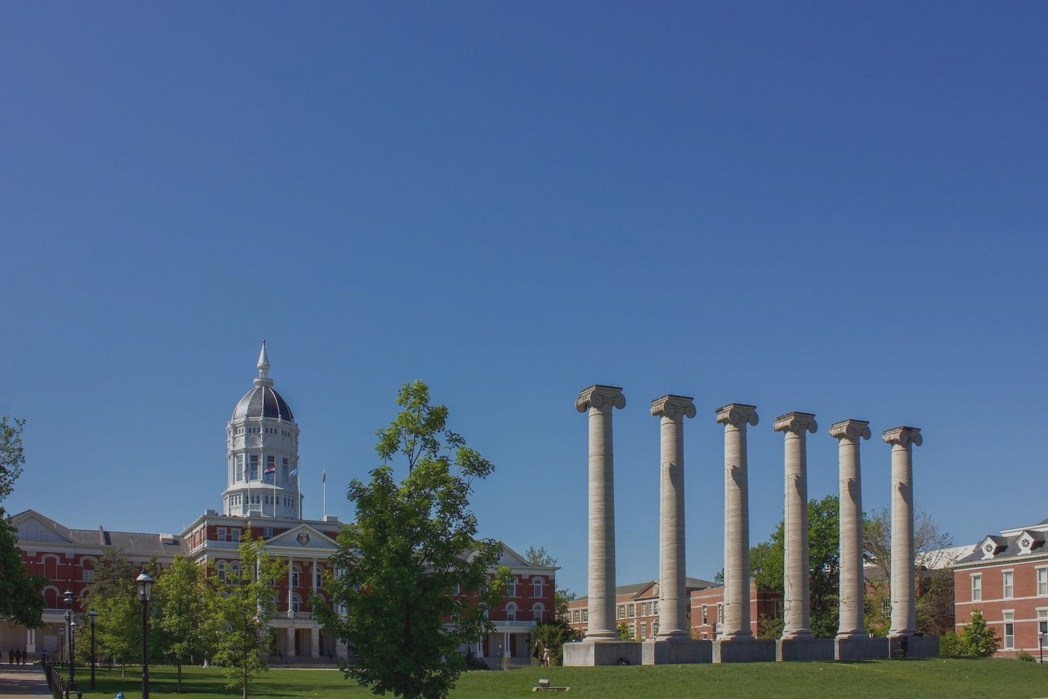 Campus lawn with a white domed building and five stone columns under a clear blue sky