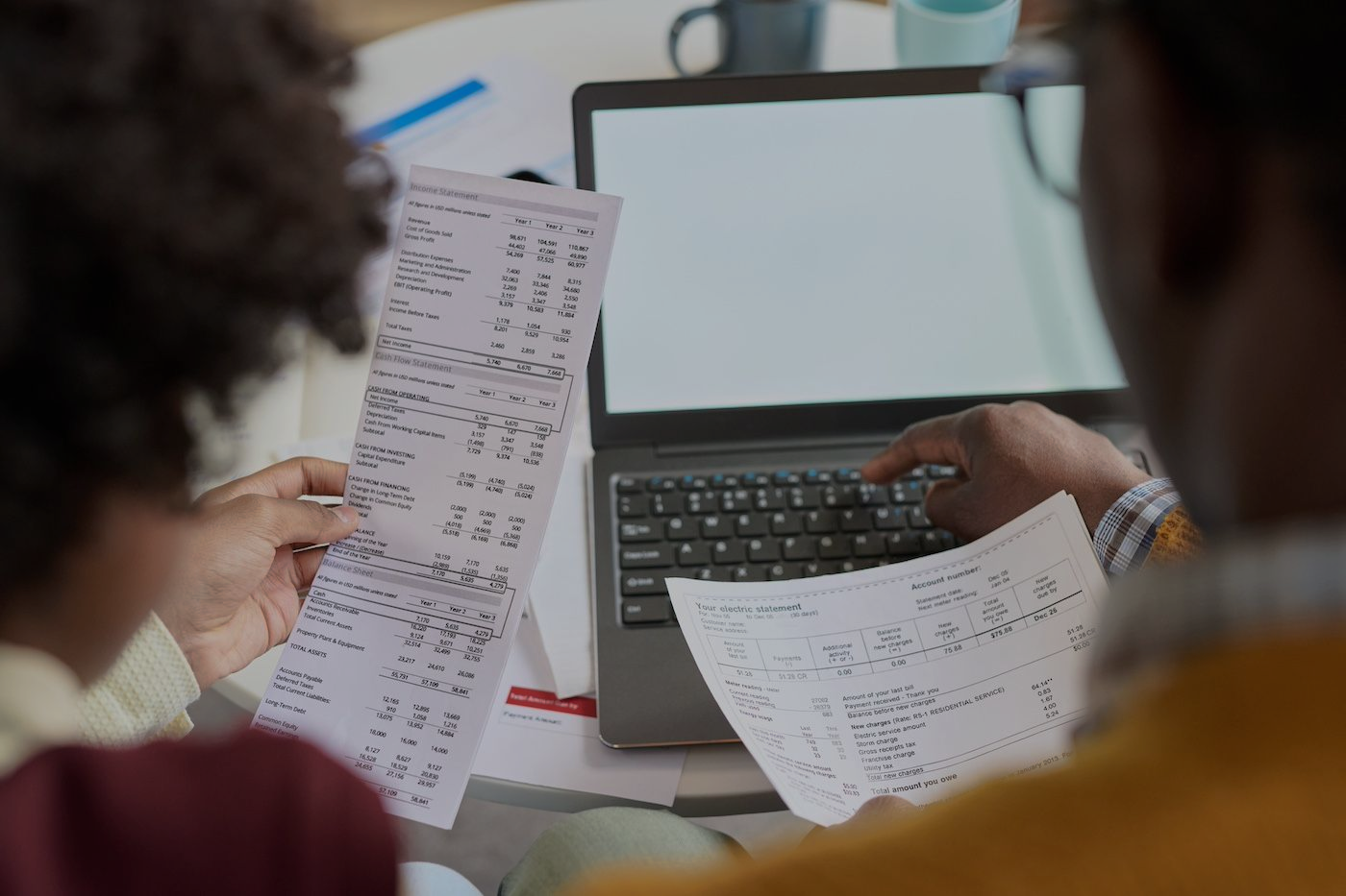 Two people examine handwritten documents while sitting at a table with a laptop open.