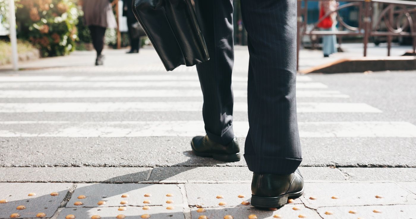 A close-up of a person’s legs and shoes as they walk across a marked pedestrian crosswalk.