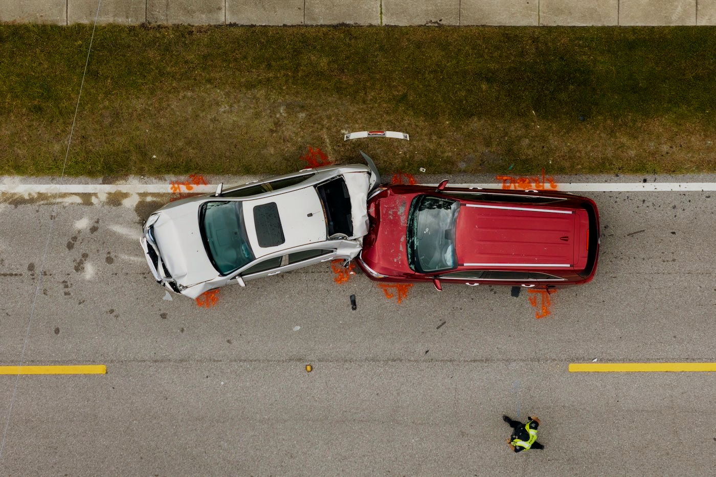 Overhead view of a car crash: a silver car and a red SUV collide on a road, with a person in a vest nearby.