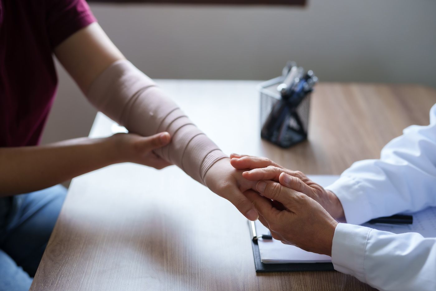 Doctor holds a patient's bandaged arm at a desk. The patient wears a burgundy shirt.