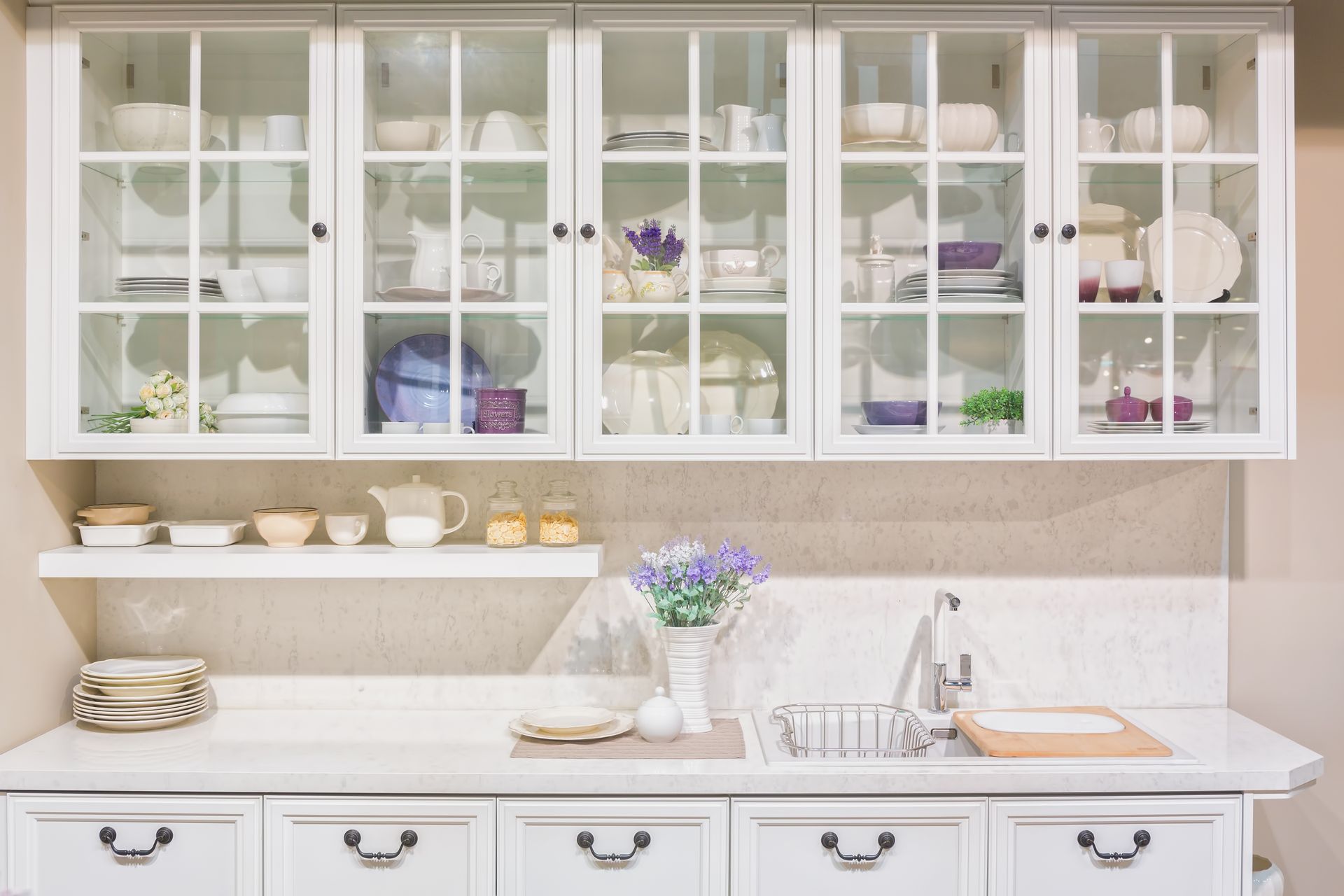 White kitchen with upper cabinets displaying dishware, countertop with sink, and lower cabinets.