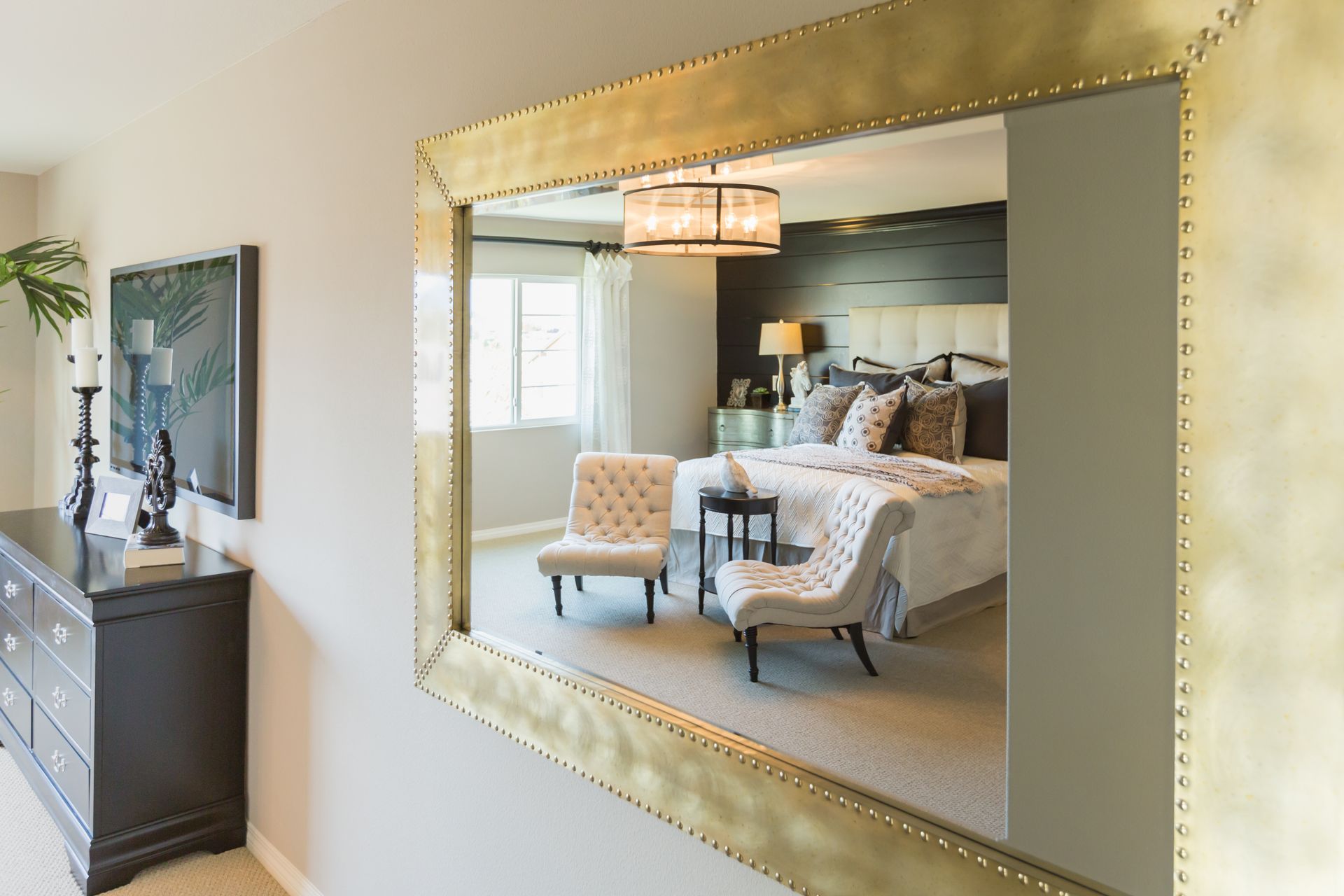 Large gold-framed mirror reflecting a bedroom with a bed, chairs, and a dresser.