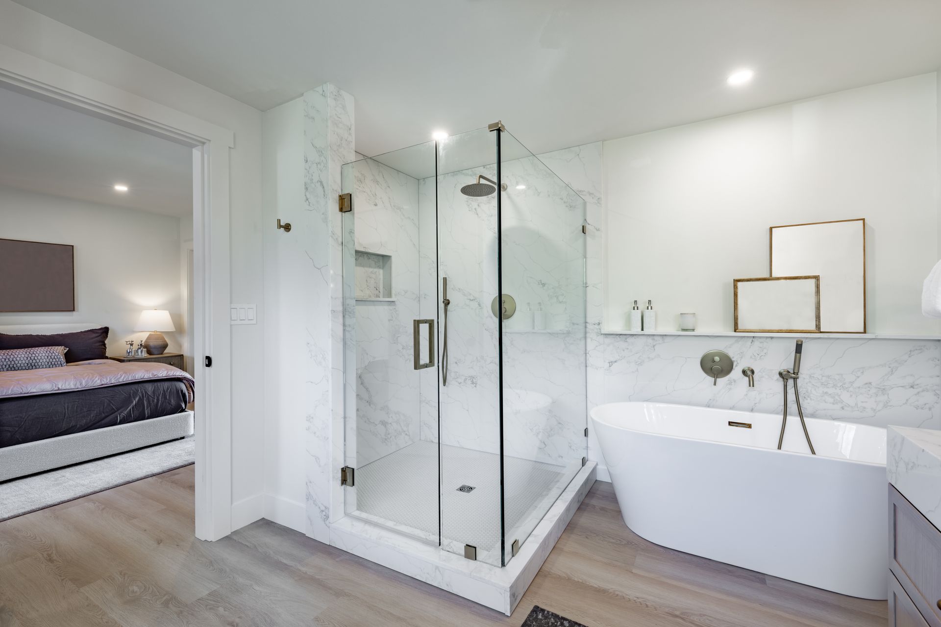 Modern bathroom with glass shower, bathtub, and view into bedroom. Light wood floors, white walls.
