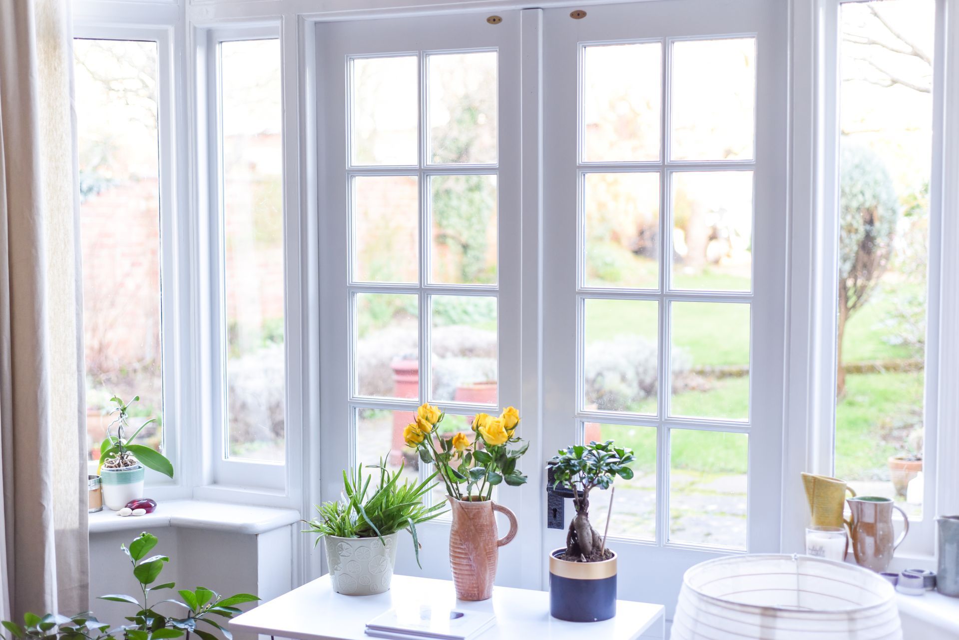 White-framed French doors and windows look out on a sunny garden. Yellow flowers and plants sit on a white table.