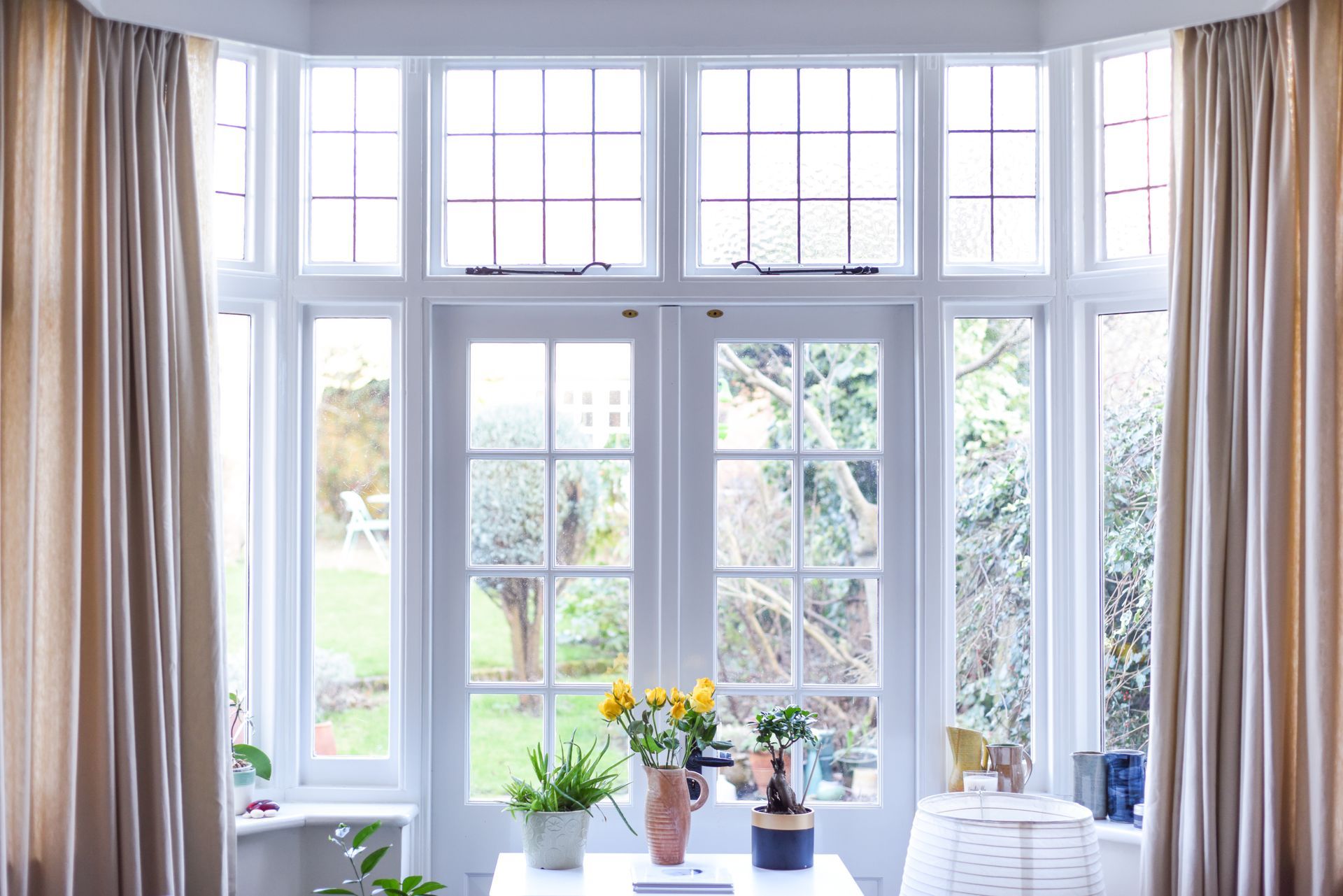 Bright white bay window overlooking a garden; cream curtains frame the view. A table has flowers and plants.