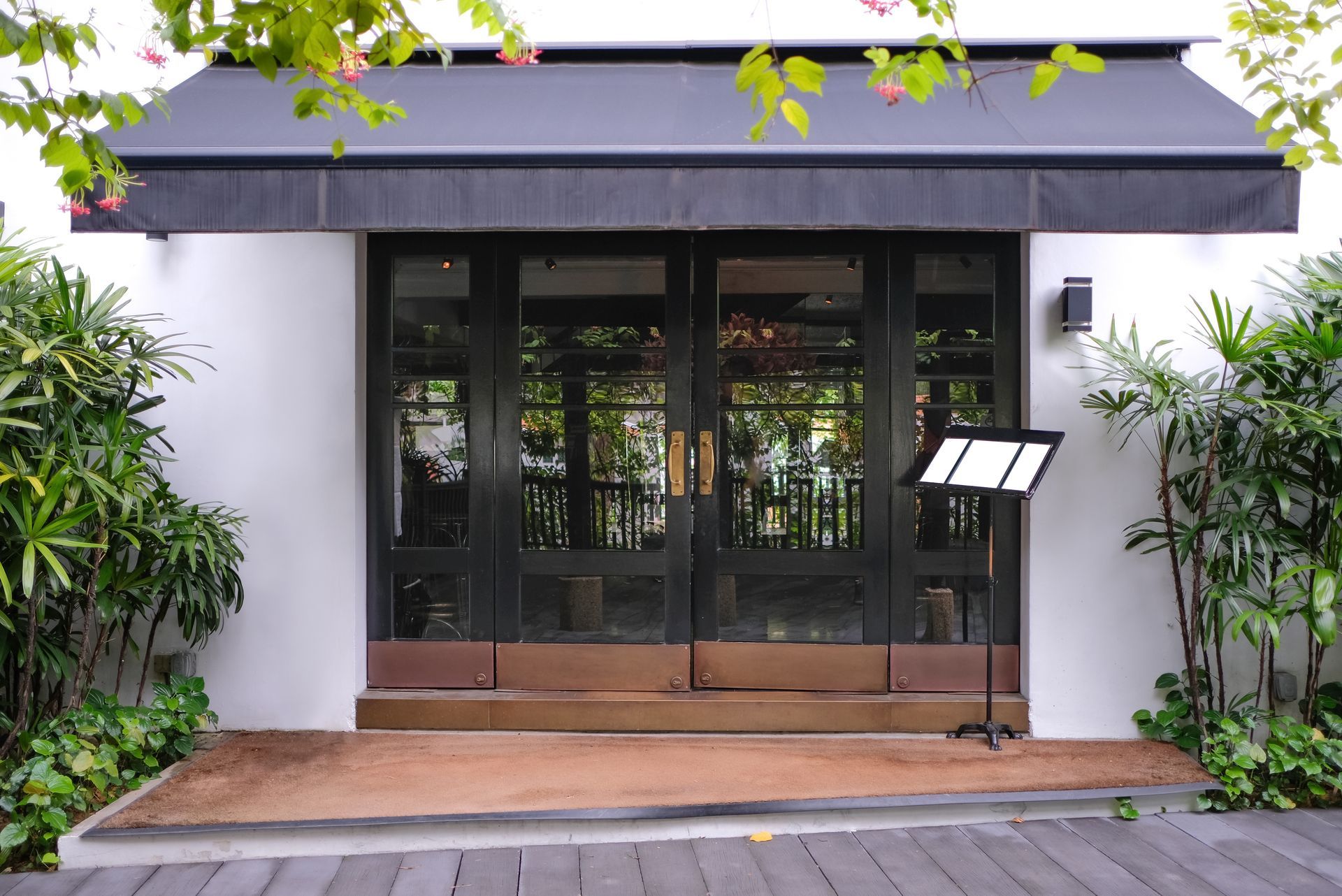Black double doors with glass panels and awning, entrance to building with greenery.
