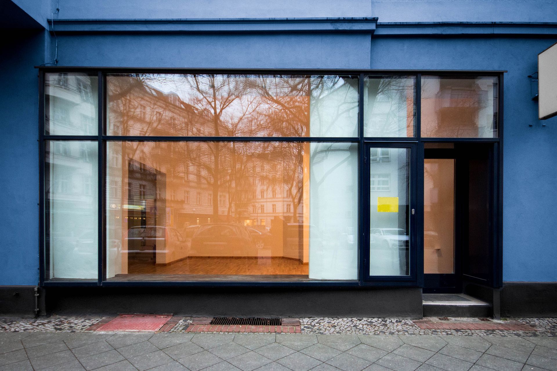 Blue building facade with large window display. Interior reflects trees with orange/brown hues. Black frame.