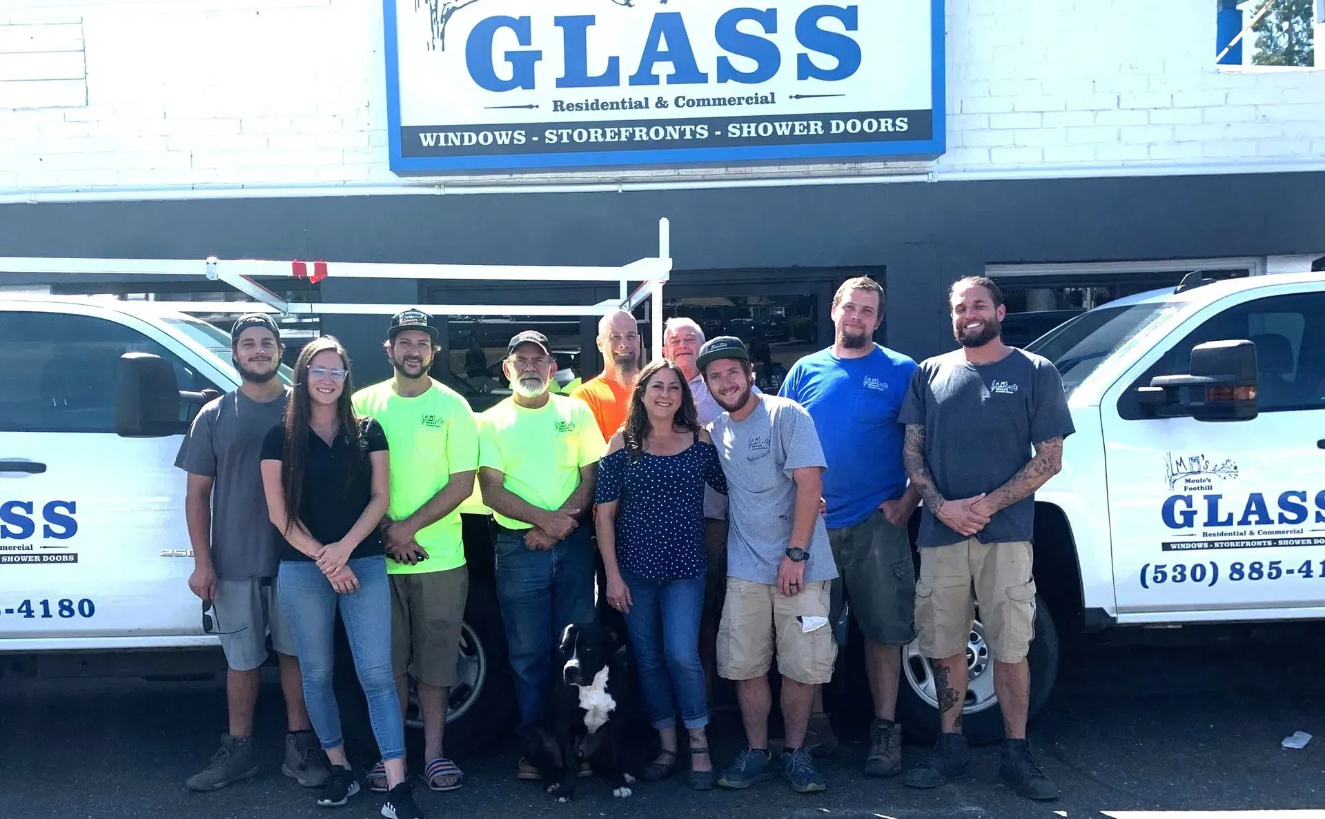 Group of people posing in front of a building with GLASS sign; two trucks with company logo.