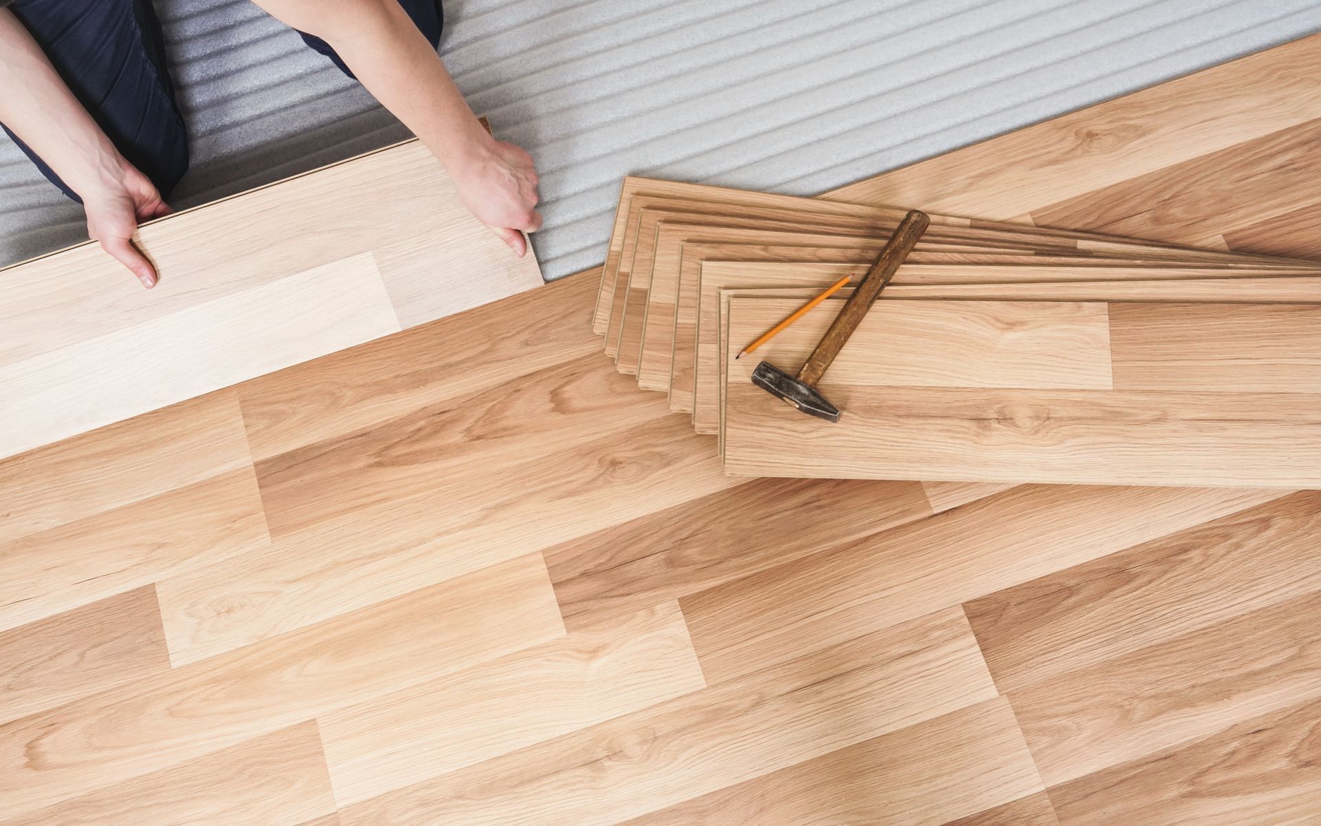 Person installing laminate flooring, hands on a plank. Floor has installed and uninstalled planks. Tools nearby.