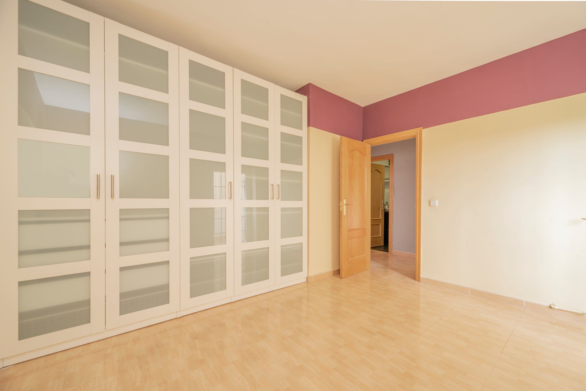 Empty bedroom with a large white wardrobe with glass doors. Light-colored wooden floor. Mauve accent wall.