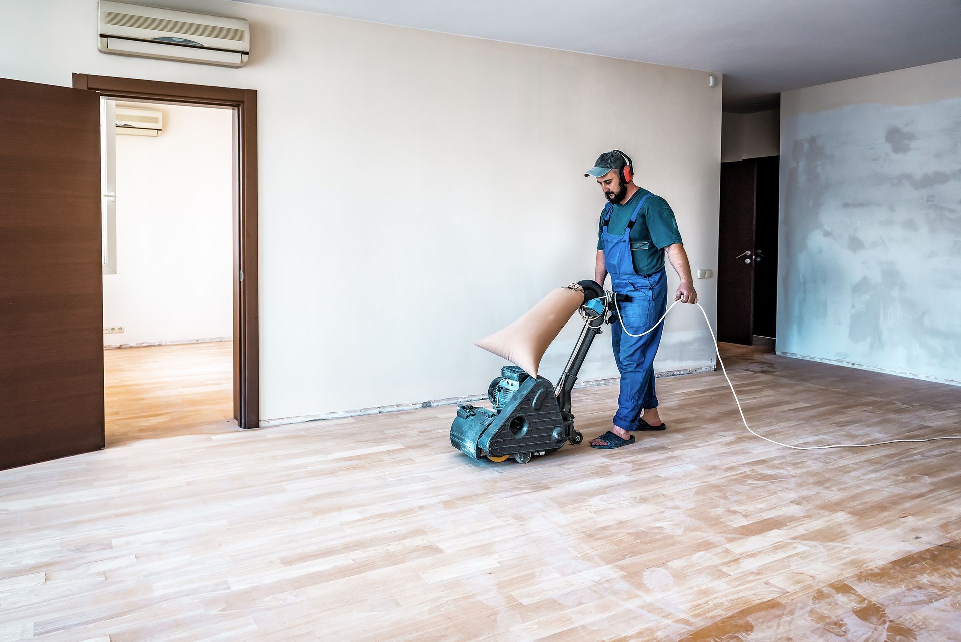 Man in blue overalls sanding hardwood floor in a room with open doorways.