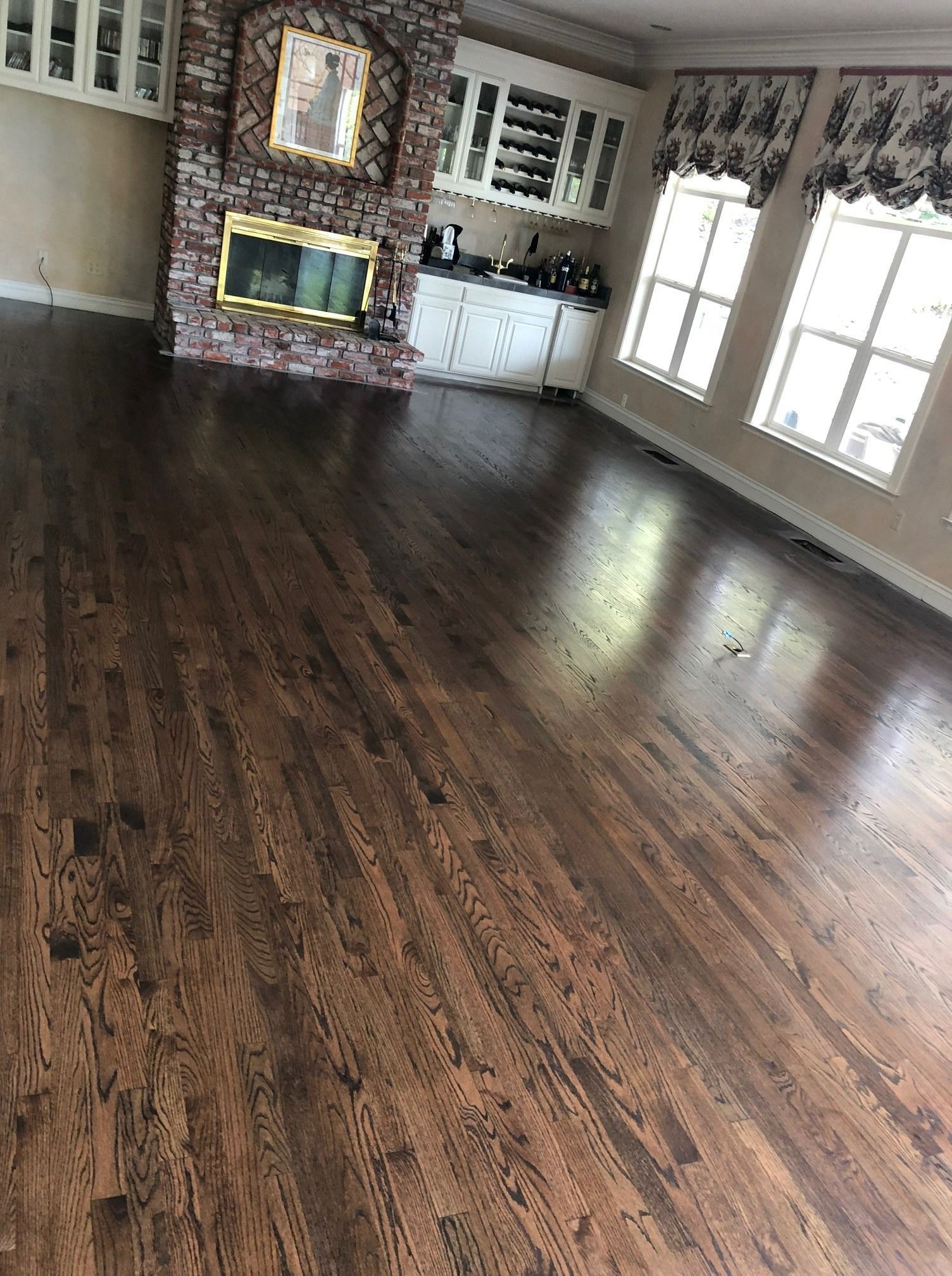 Dark stained hardwood floor in a room with a brick fireplace, built-in shelves, and windows with curtains.