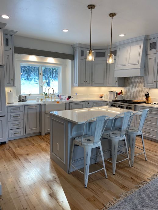 A modern kitchen with light gray distressed cabinets, a white marble island with three metal stools, and hardwood floors.