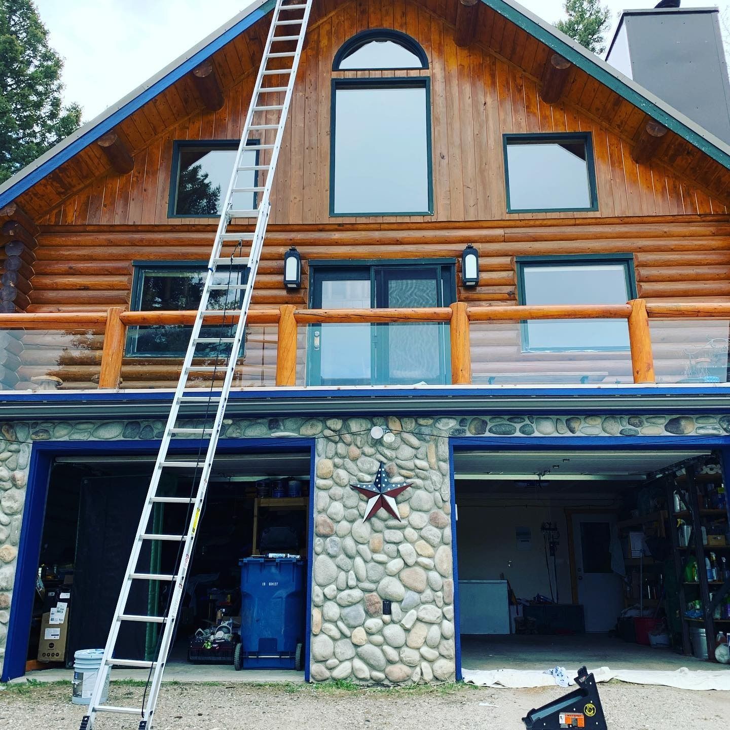 An extension ladder rests against the front facade of a two-story log cabin home with a stone-covered garage base.