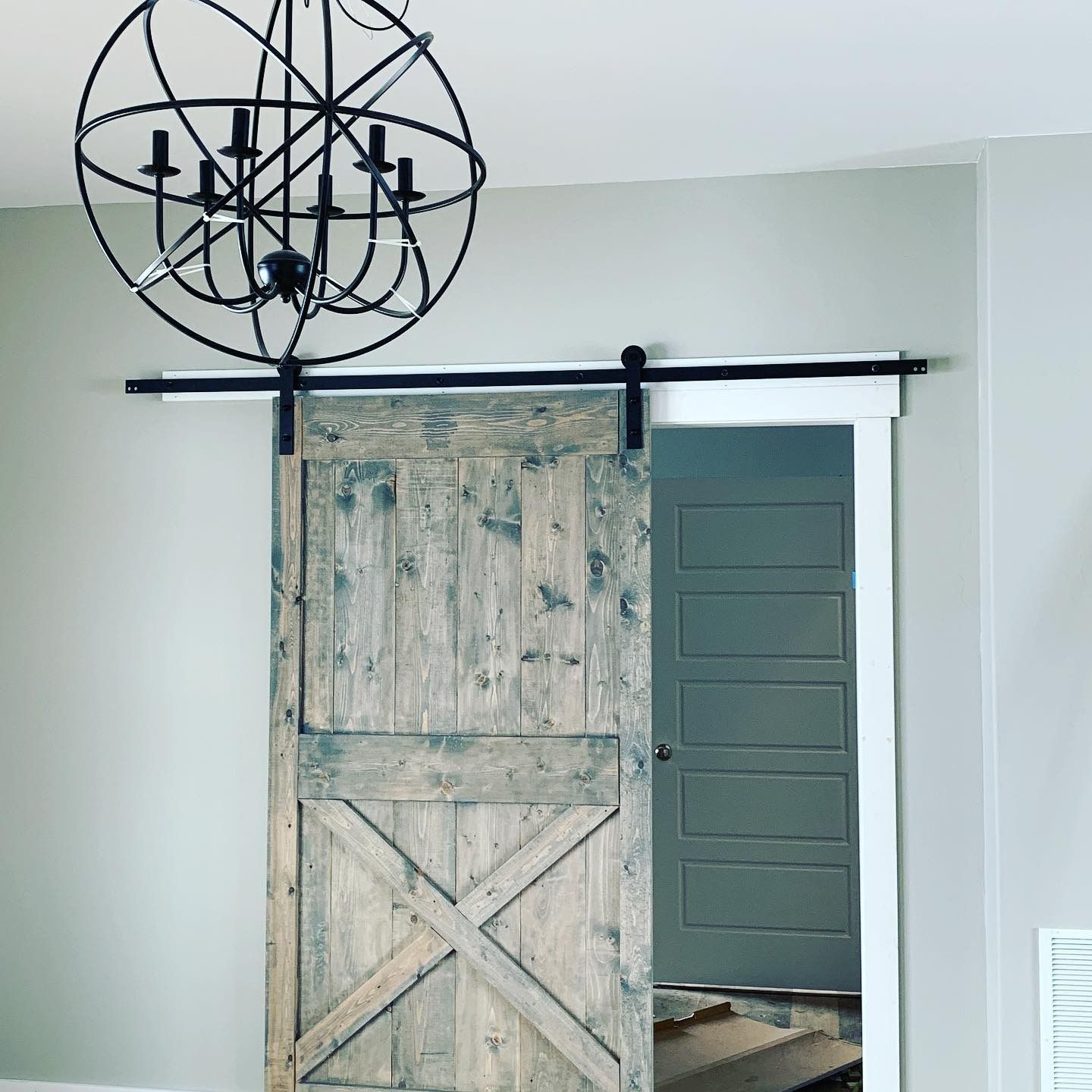 A rustic wood barn door with an X-brace design hangs on a black metal track before a gray interior doorway and orb light.