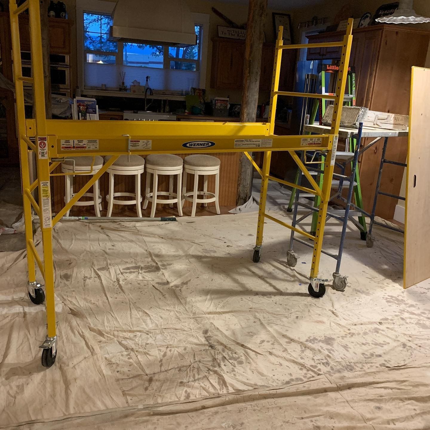 A yellow metal rolling scaffold stands on a drop cloth in a home interior with a kitchen island and bar stools in the back.